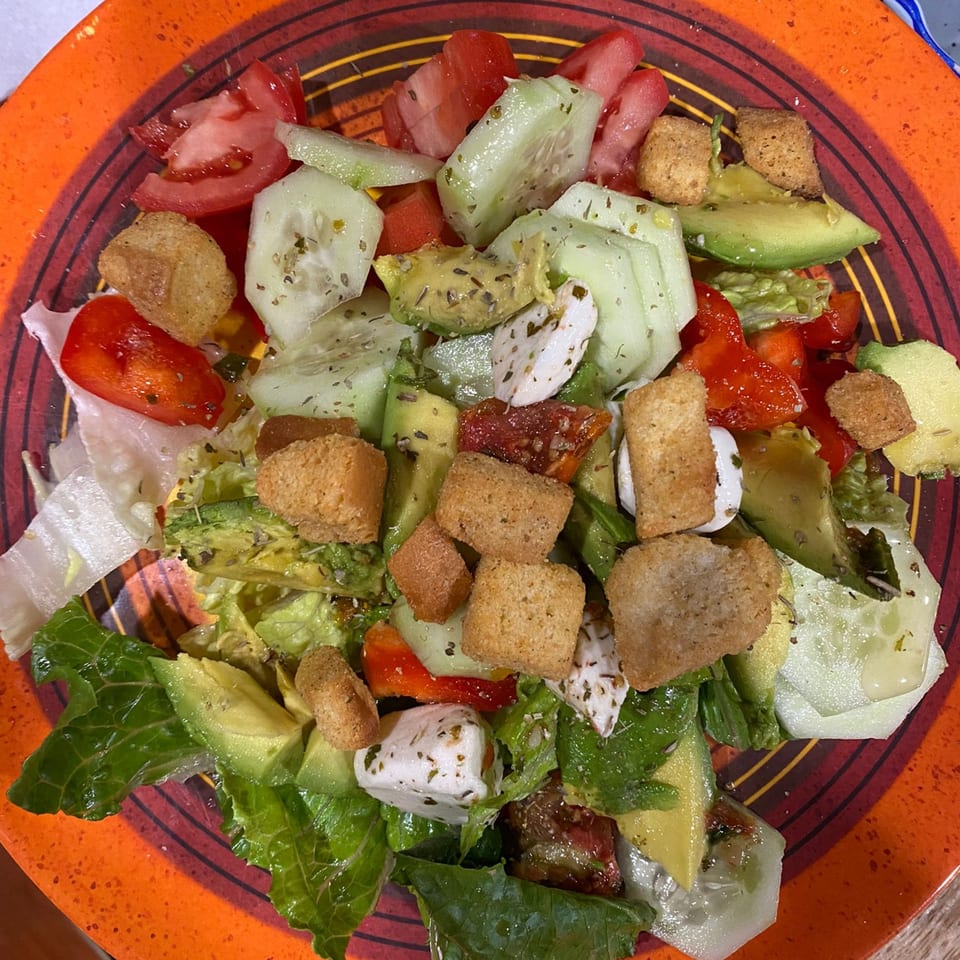 Vibrant salad with lettuce, tomatoes, cucumbers, avocado, croutons, and feta cheese on an orange plate.
