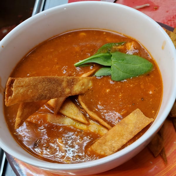 A white bowl of rich red tomato soup with tortilla strips and fresh green basil.