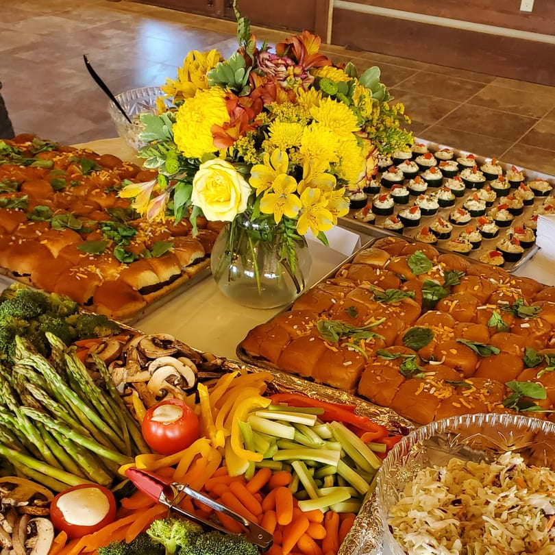 A buffet table laden with sliders, vegetable platters, and small appetizers, adorned with a yellow floral centerpiece.