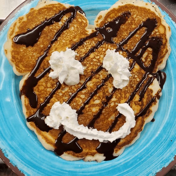 Mickey Mouse-shaped pancakes with chocolate syrup and whipped cream on a blue plate.