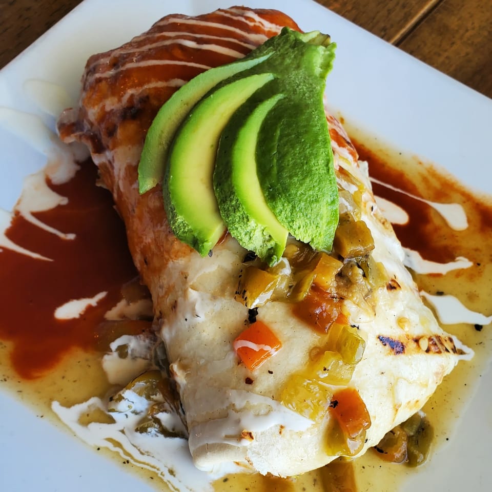 A delicious burrito topped with sliced avocado, green chile, and red and white sauces.