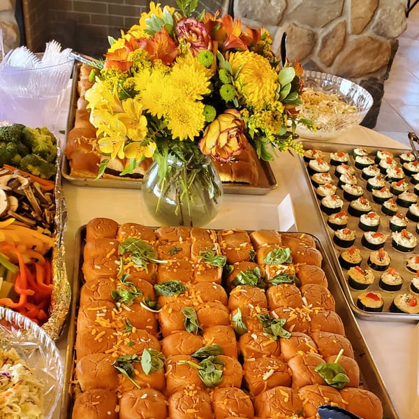 A buffet table with sliders, vegetable platters, and appetizers, adorned with a vibrant floral centerpiece.