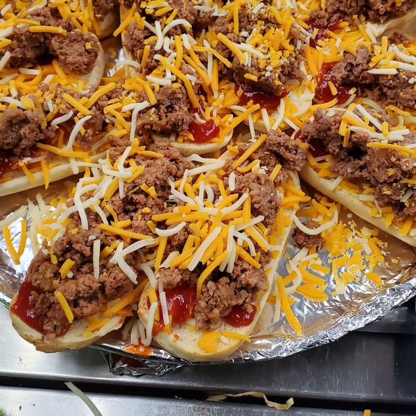 Close-up of mini hot dog buns topped with ground meat, shredded cheese, and ketchup.