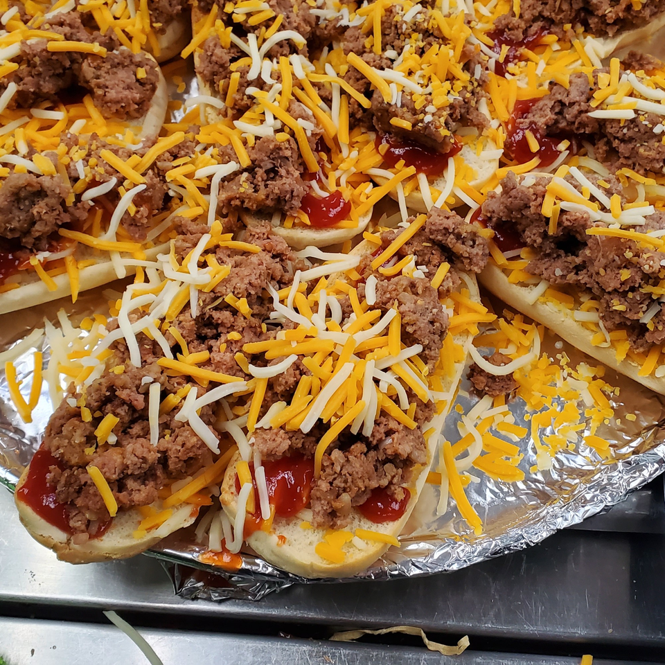 Close-up of mini hot dog buns topped with ground meat, shredded cheese, and ketchup.